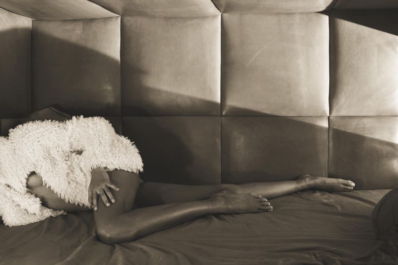 Model lying on a bed wearing a fluffy white textured coat against a paneled wall.