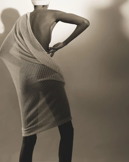 Woman in stylish pose wearing a draped knitted dress and head wrap, back view, against a plain background in a studio setting.