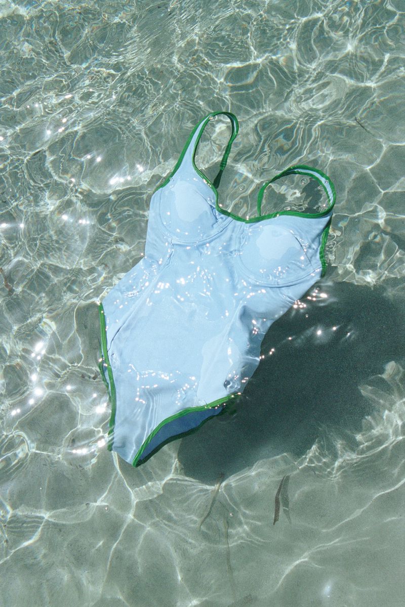 Light blue one-piece swimsuit with green trim submerged in shallow water, creating shimmering light patterns on the fabric and surrounding sand.
