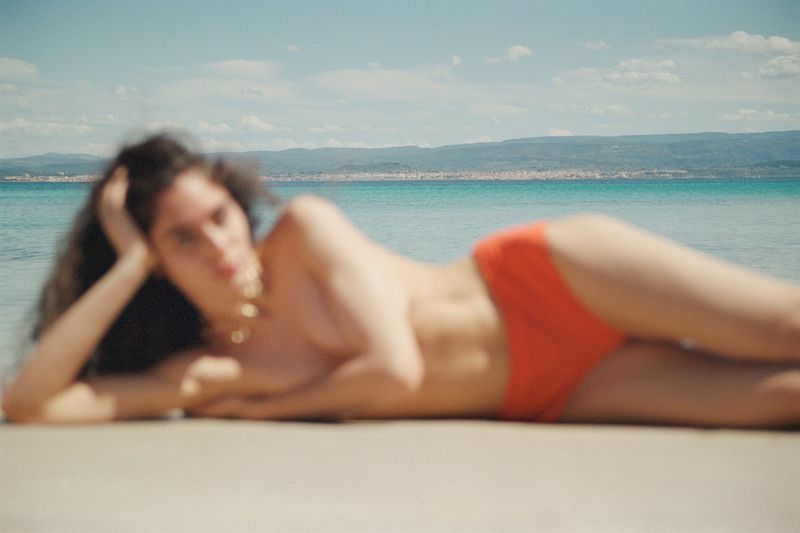 Brunette woman in swimwear lounges on sandy beach with turquoise water and island horizon under a bright blue sky with clouds.