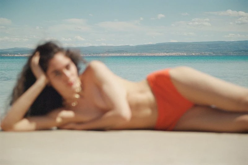 Brunette woman in swimwear lounges on sandy beach with turquoise water and island horizon under a bright blue sky with clouds.