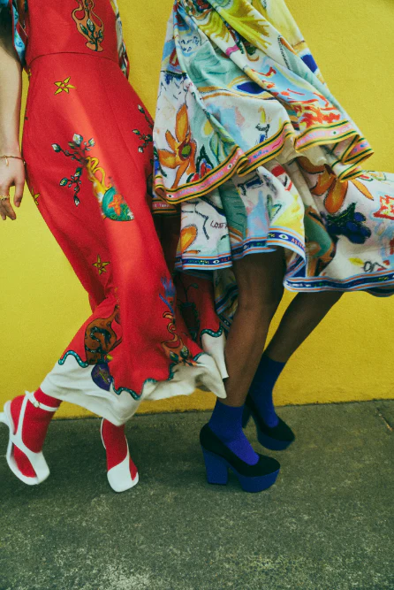 Two women stand in colorful dresses and platform heels against a yellow wall, showcasing a fashion-forward and vibrant editorial shot.