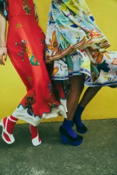 Two models wearing patterned dresses and platform shoes against a bright yellow wall and concrete ground for Alemais