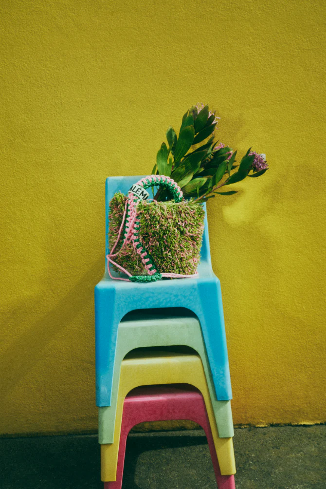 Moss-covered handbag filled with flowers on stacked colorful chairs against a yellow wall, showcasing a blend of nature and design.