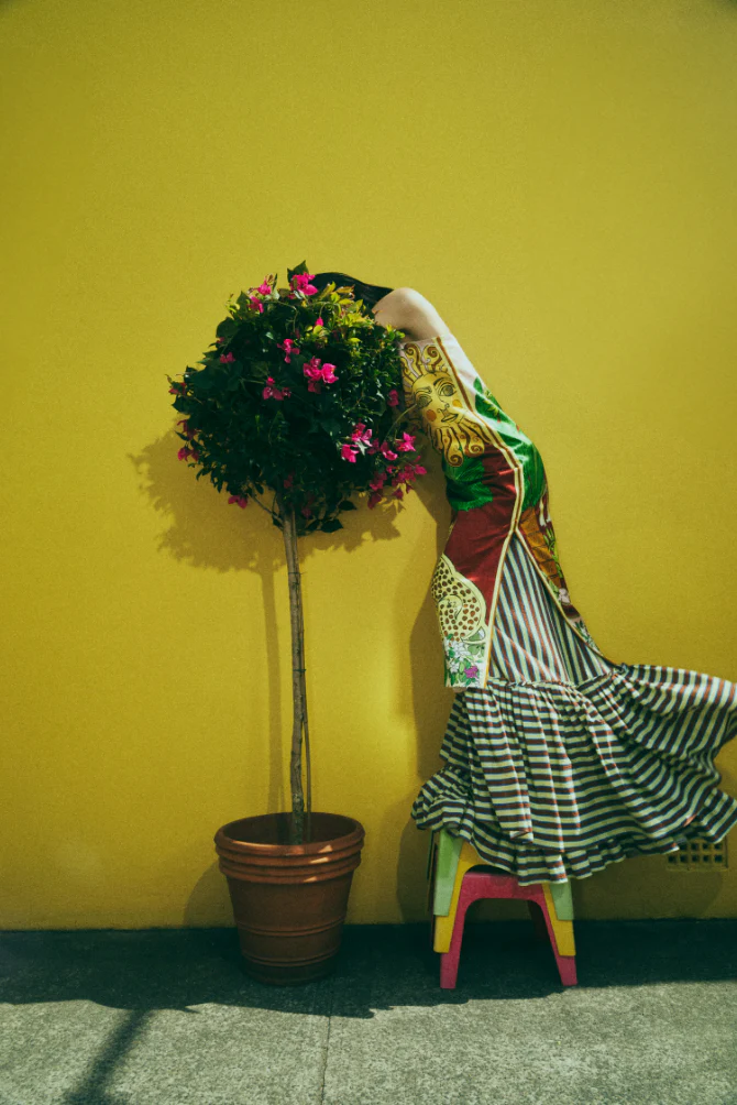 Woman in colorful dress leans against yellow wall next to potted plant with pink flowers in outdoor setting on city street.