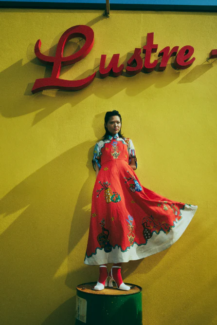 Young woman in vibrant red dress and boots standing on a metal barrel in front of a yellow wall with a red 'Lustre' sign.