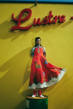 Young woman in vibrant red dress and boots standing on a metal barrel in front of a yellow wall with a red 'Lustre' sign.