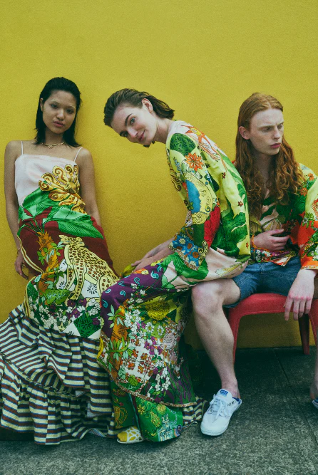 Three fashion models pose against a yellow wall wearing colorful, patterned clothing and white sneakers, set outdoors in natural light.