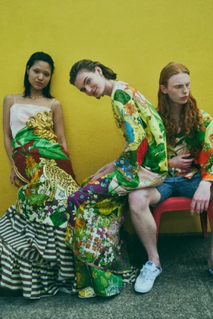Three fashion models pose against a yellow wall wearing colorful, patterned clothing and white sneakers, set outdoors in natural light.