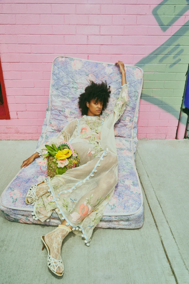 A woman in a floral dress poses on a mattress in an outdoor setting with a pink brick wall and floral basket in a fashion-forward image.