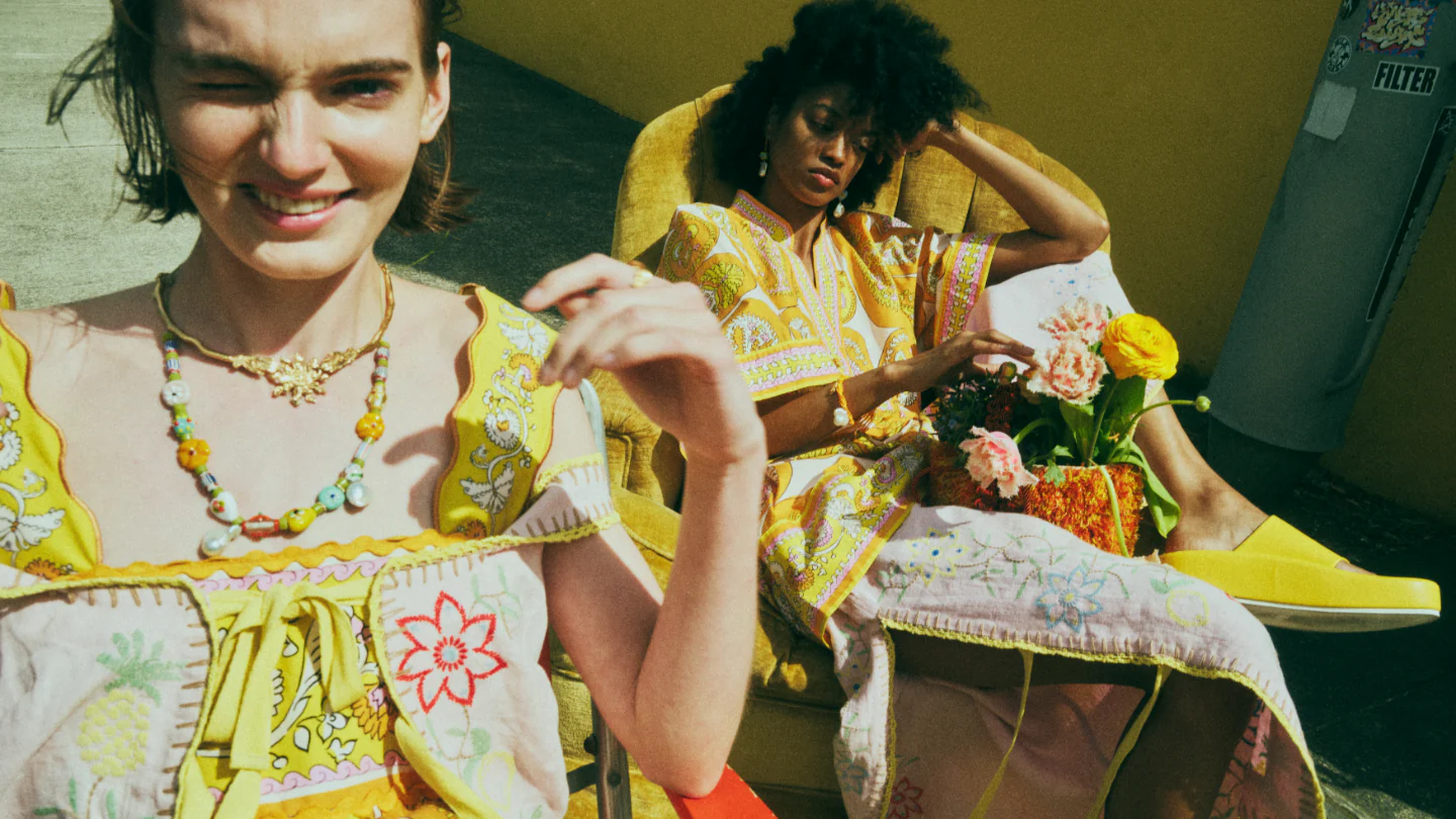 Two women in patterned yellow outfits pose outdoors, one winking, the other reclining with flowers, creating a summery, vintage fashion scene.