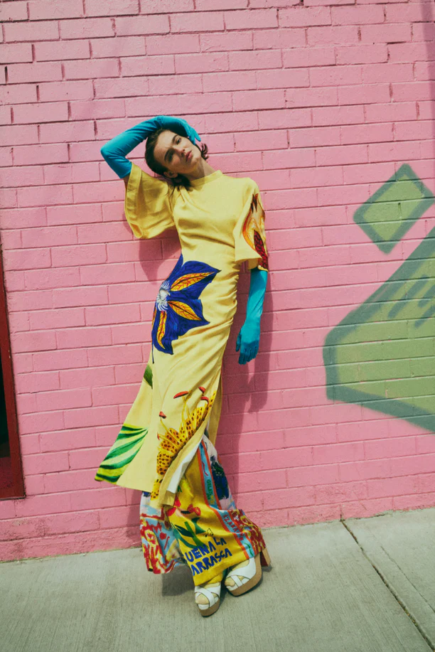 A woman in a yellow patterned dress poses against a pink brick wall, wearing blue gloves and platform sandals in an outdoor shot.