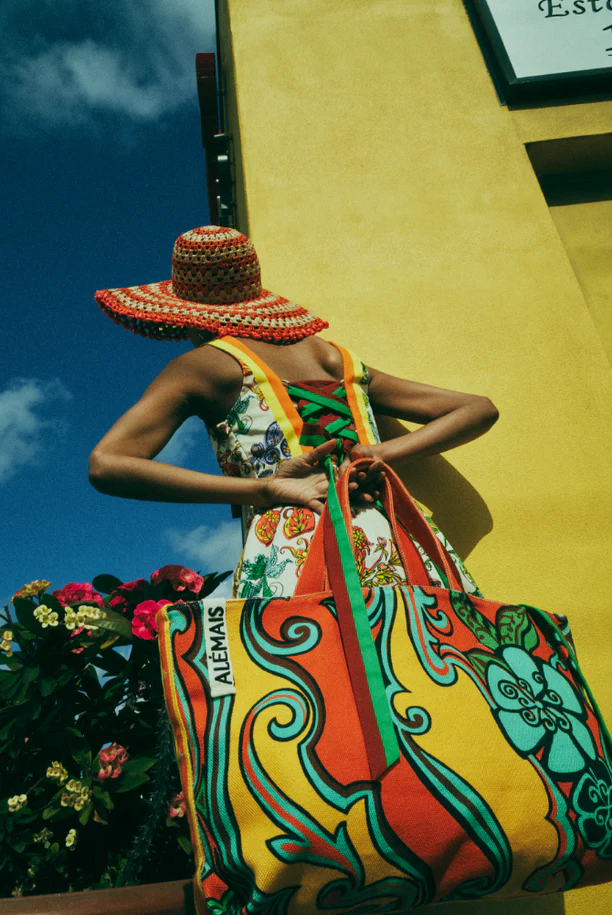 A woman models a colorful tote bag and sun hat against a bright yellow building and blue sky, showcasing summer fashion and style.