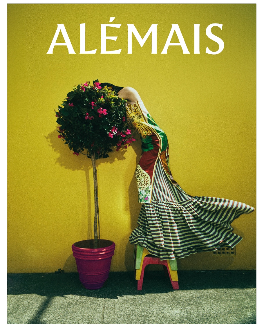 Fashion image featuring a model in a colorful dress bending over a potted plant, with a yellow wall and a small stool in the background.