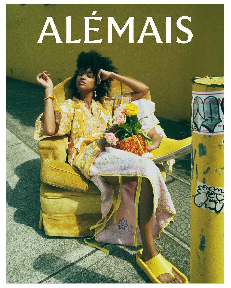 Black woman in yellow floral dress and slides sits in a yellow armchair on concrete with a bouquet of flowers in dappled sunlight.