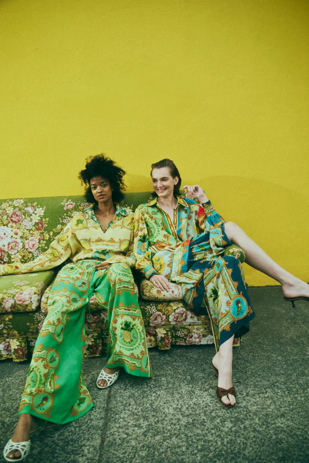 Two women pose in patterned matching sets on a floral couch against a yellow wall. They wear trendy patterned tops and bottoms.