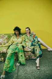 Two female models wearing patterned green and yellow matching outfits seated on a floral patterned sofa against a yellow wall.