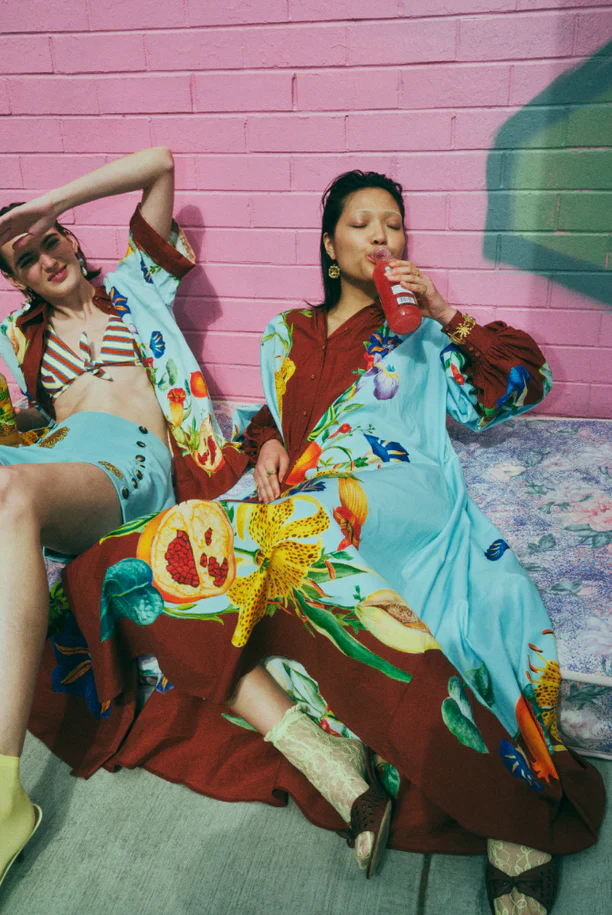 Two women in patterned outfits sit against a pink brick wall, one drinking from a bottle, showcasing fashion and vibrant colors.