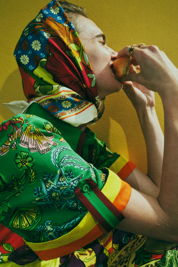 Adult wearing colorful printed shirt and headscarf eats orange against a yellow backdrop in a fashion-style editorial portrait.
