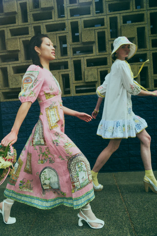 Two women walk in front of a textured building. The woman in the foreground is wearing a pink dress with white shoes and carrying a bouquet.