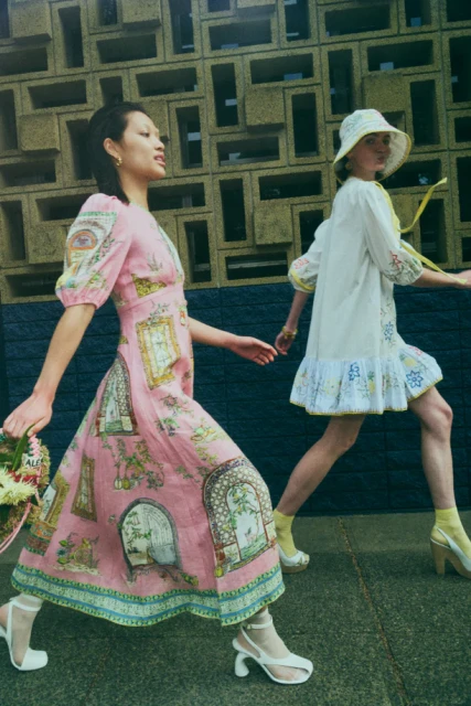 Two women walk in front of a textured building. The woman in the foreground is wearing a pink dress with white shoes and carrying a bouquet.