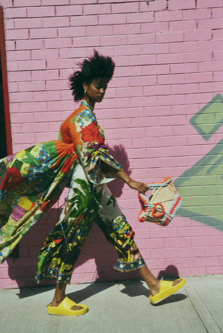 Woman walking in vibrant printed outfit and yellow slides against a textured pink brick wall, carrying a patterned purse. Fashion forward.