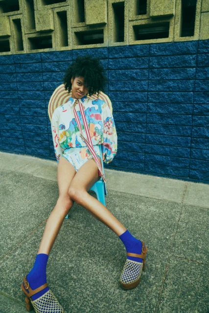 A young woman with curly hair poses seated outdoors in a colorful patterned outfit, blue socks, and clogs, against a textured blue wall.