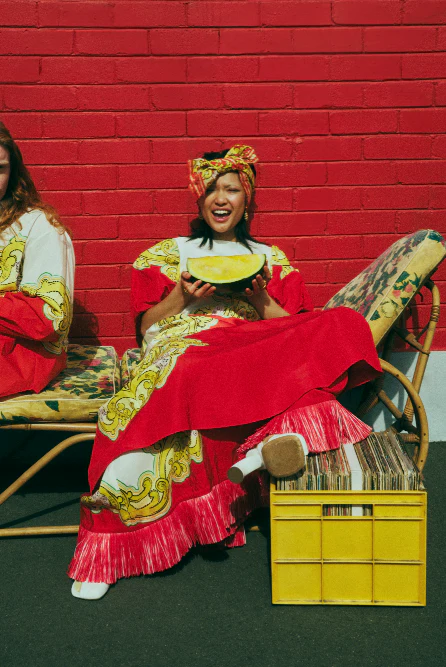 Two women in patterned dresses sit on floral chairs with red brick background. One woman holds a slice of watermelon in her hands.