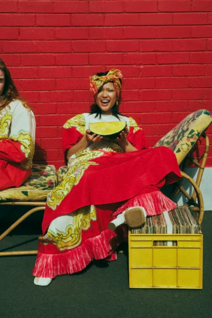 Two women in patterned dresses sit on floral chairs with red brick background. One woman holds a slice of watermelon in her hands.