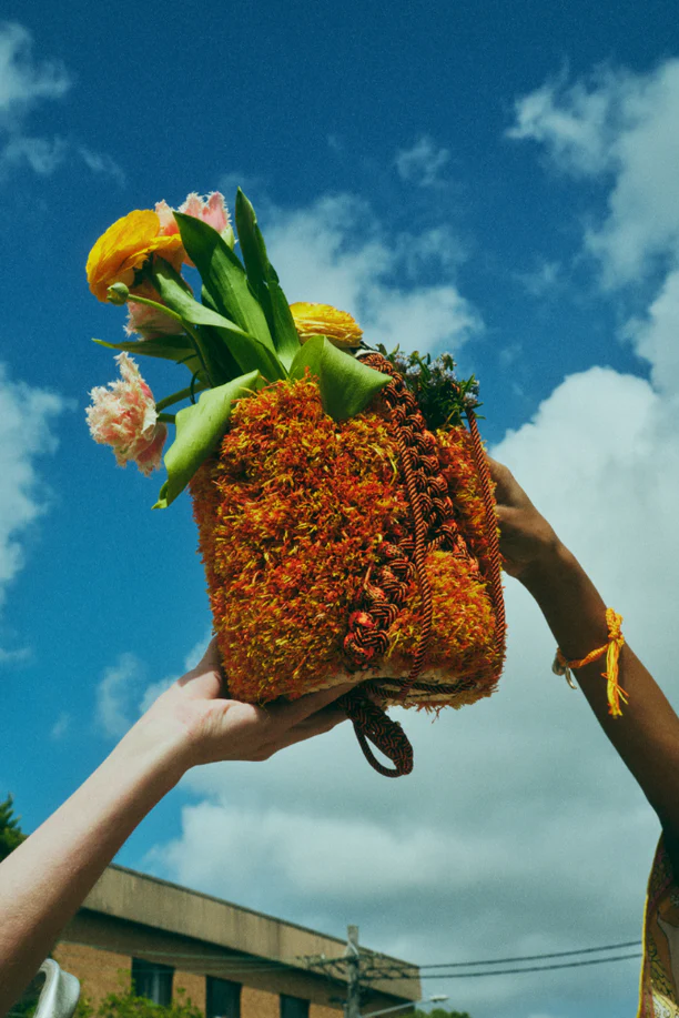 Floral arrangement in an orange textured bag held up against a cloudy blue sky by two people, one with a yellow bracelet.