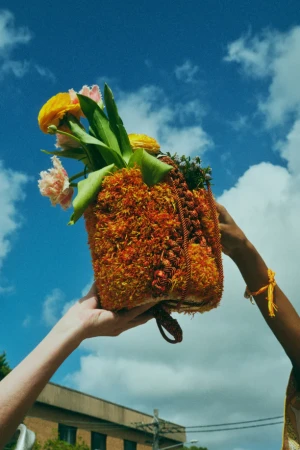 Floral arrangement in an orange textured bag held up against a cloudy blue sky by two people, one with a yellow bracelet.