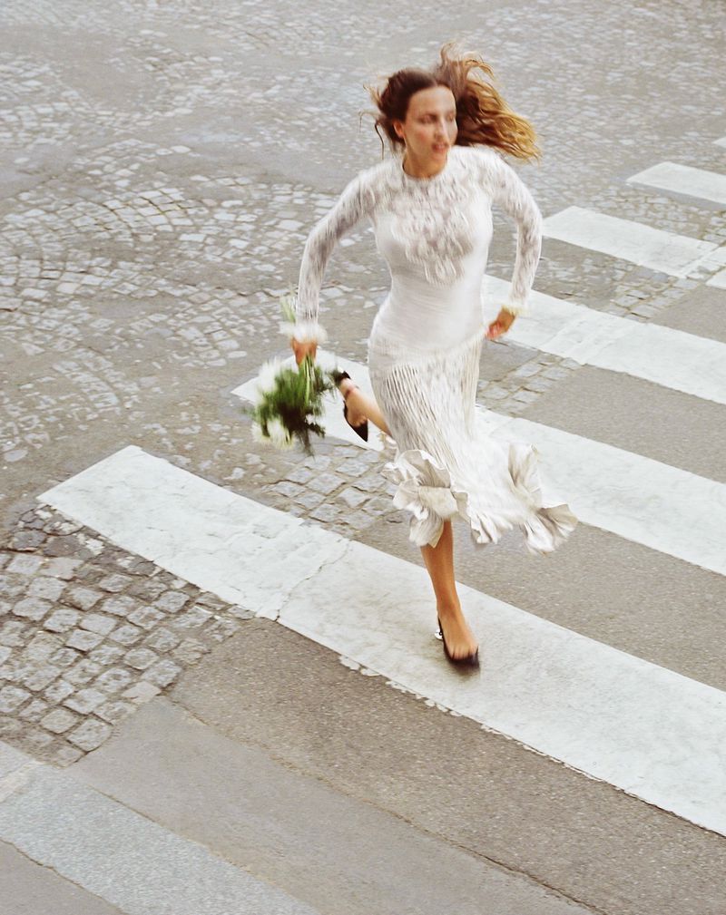 Woman in long-sleeved white dress and dark heels sprints across a crosswalk holding white flowers on a cobblestone road.