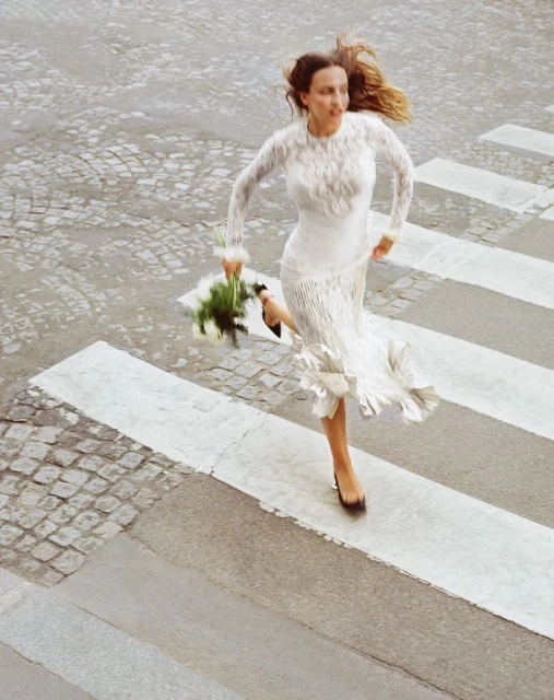 Woman in long-sleeved white dress and dark heels sprints across a crosswalk holding white flowers on a cobblestone road.