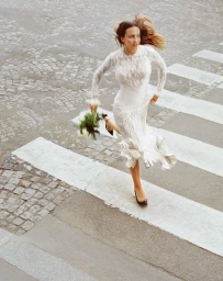 Model in white lace wedding dress running across a cobbled street with a bouquet of flowers for Rabanne