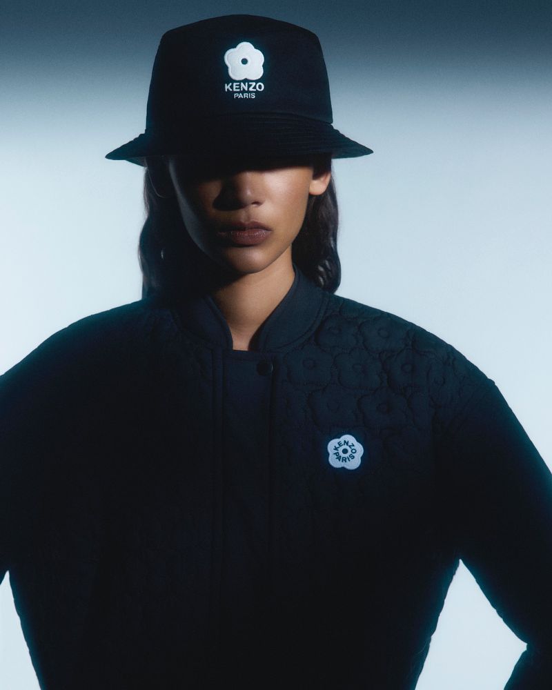 Portrait of a woman wearing a black patterned bomber jacket and a Kenzo Paris bucket hat with a floral logo against a light blue backdrop.