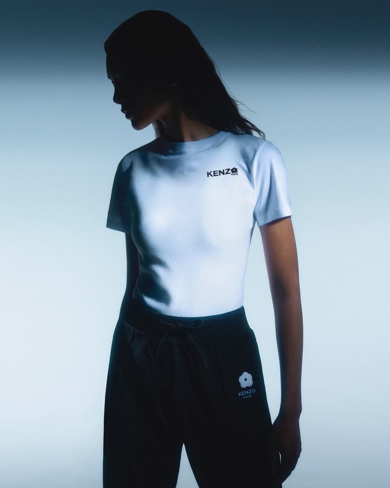 A woman posing in a white Kenzo t-shirt and dark sweatpants, set against a gradient blue and white backdrop.