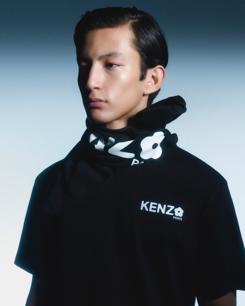 Portrait of a young adult male wearing a black Kenzo t-shirt with a white floral design and logo, against a gradient blue background.