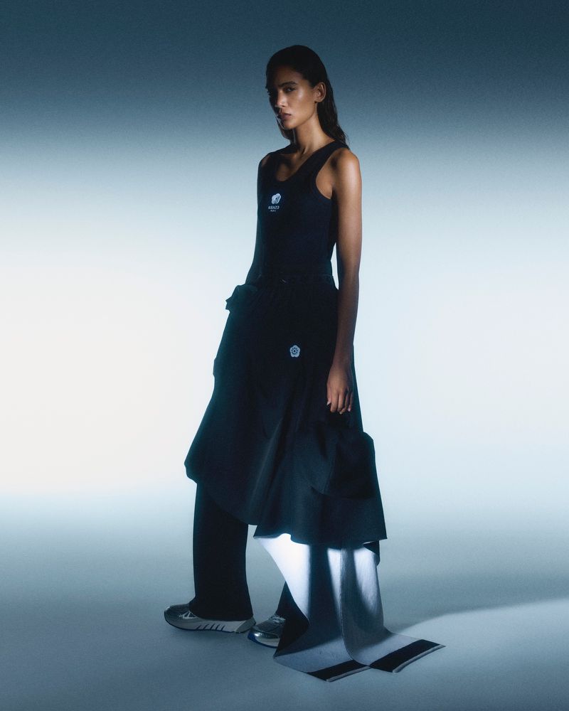 A woman in a sleeveless top, pants, and skirt poses indoors with soft studio lighting, creating a fashion editorial image with subtle color grading.