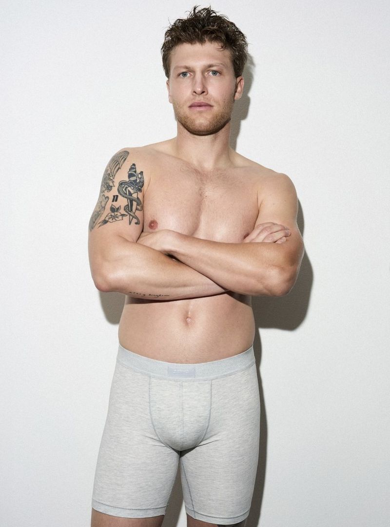 Adult male posing shirtless in boxer briefs against a white background, wearing boxer briefs and displaying arm tattoos.