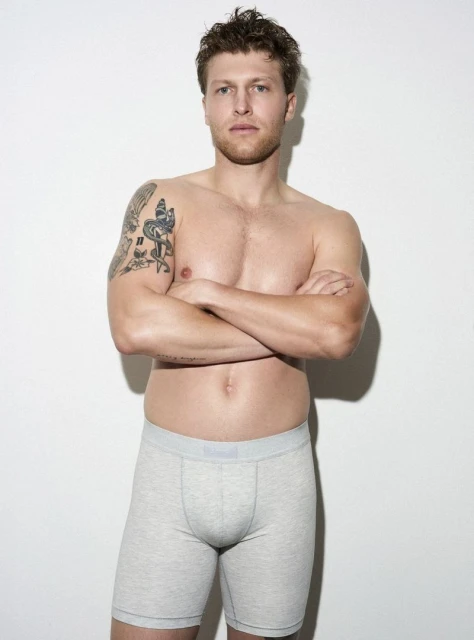 Adult male posing shirtless in boxer briefs against a white background, wearing boxer briefs and displaying arm tattoos.