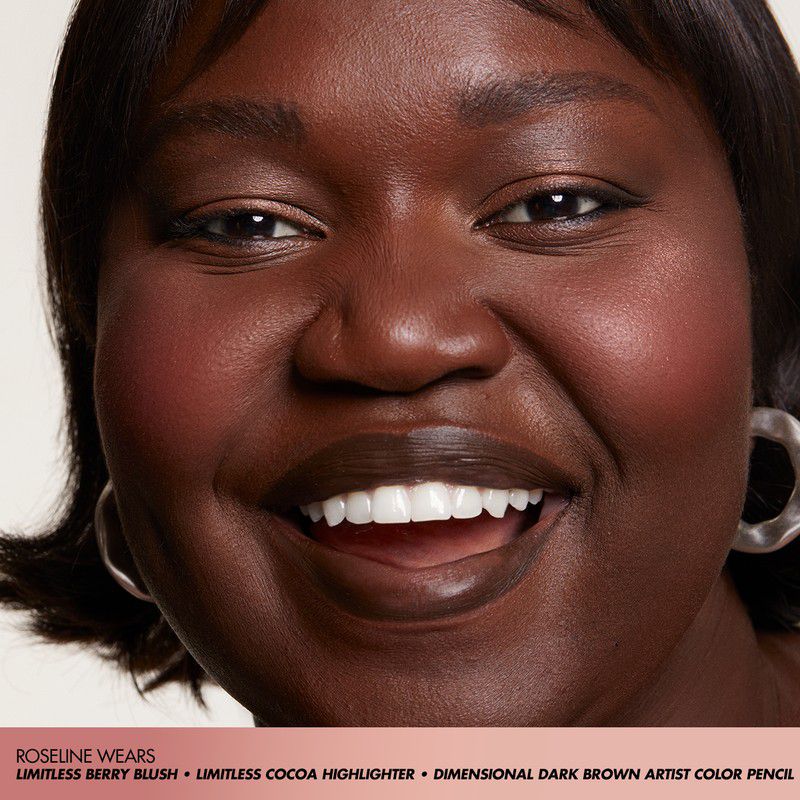 Close-up portrait of an African-American woman with dark skin smiling, wearing blush, highlighter, and earrings against a light background.