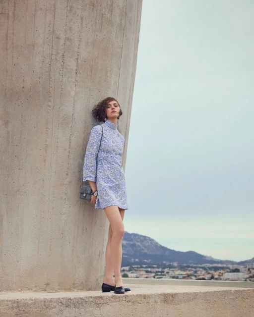 Adult woman wearing a lilac mini dress with a purse posing against a concrete structure with a city and mountain view in the background.