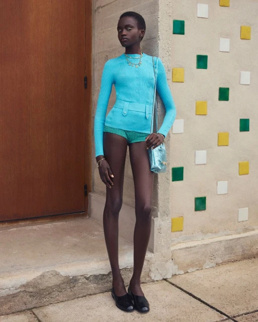 A South Sudanese fashion model poses in a light-blue knitwear outfit, accented with a shiny blue purse, against a colorful block wall.