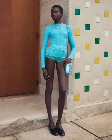 A South Sudanese fashion model poses in a light-blue knitwear outfit, accented with a shiny blue purse, against a colorful block wall.