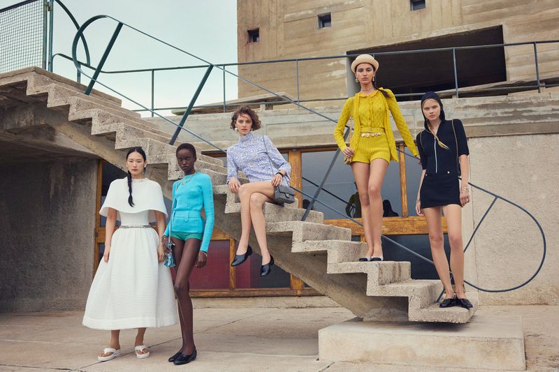 Five fashion models pose in coordinated outfits on concrete steps outside a modern building under a cloudy sky. 