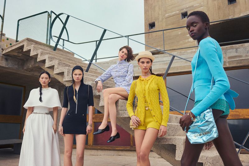 Five diverse women in a fashion editorial on concrete steps, wearing colorful coordinated outfits with purses, jewelry, and hats, set outdoors.