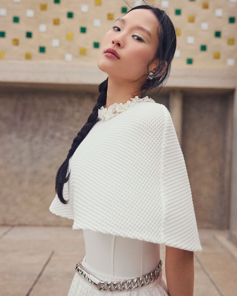 Woman with braided hair in a white textured cape stands against a mosaic tile wall, showcasing fashion and beauty in an outdoor setting.