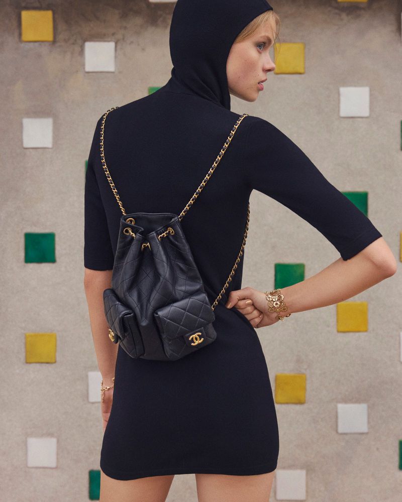 Model wearing a black hooded dress and black leather backpack with gold chain straps against a textured tiled wall.