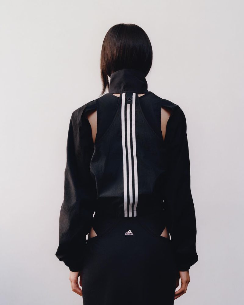 Back view of an adult woman with short dark hair wearing a black Adidas jacket with white stripes and matching black skirt against a white background.