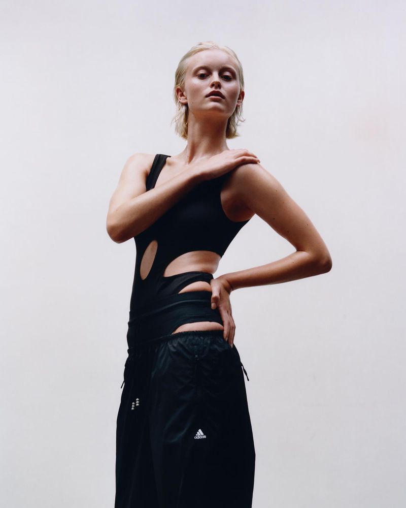 Blond-haired model poses in a black cut-out bodysuit and track pants against a white background in a studio portrait. Fashion.